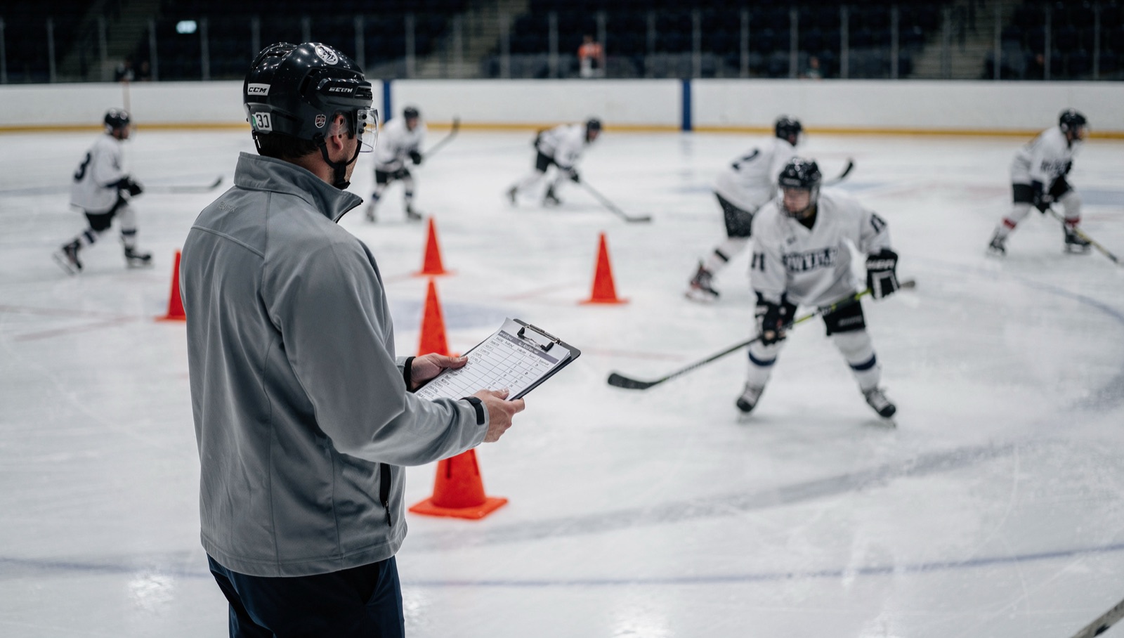 A coach with a clipboard at the boards watches young skaters in numbered jerseys do a crossover drill around pylons